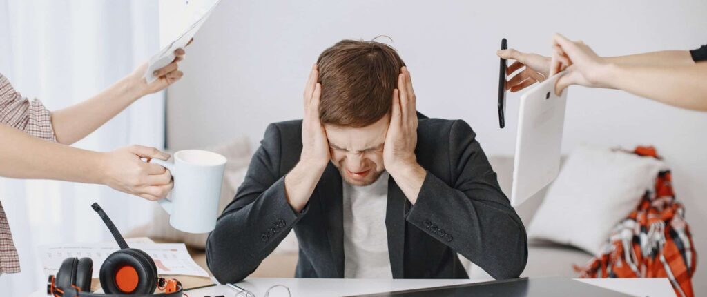 seorang pria menundukkan kepala sambil menutup telinga dikelilingi oleh kertas, ponsel, tablet, dan laptop seorang pria menundukkan kepala sambil menutup telinga dikelilingi oleh kertas, ponsel, tablet, dan laptop