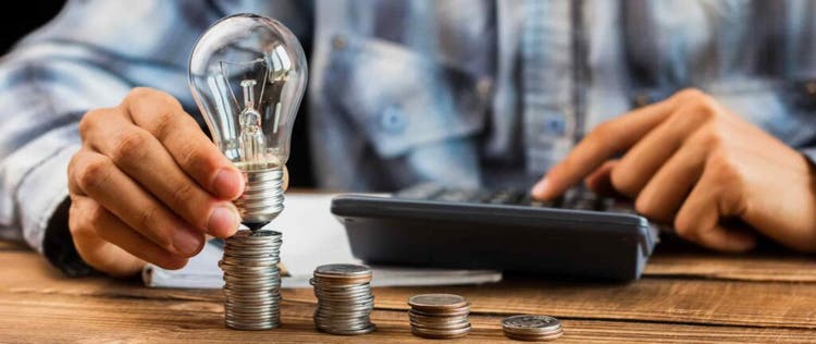 seseorang sedang menghitung sesuatu di kalkulator dan memegang bola lampu di atas tumpukan koin
