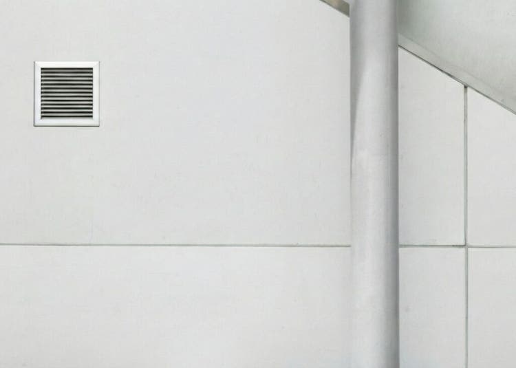 Dinding putih minimalis dengan lubang ventilasi kecil di kiri dan tiang vertikal di kanan