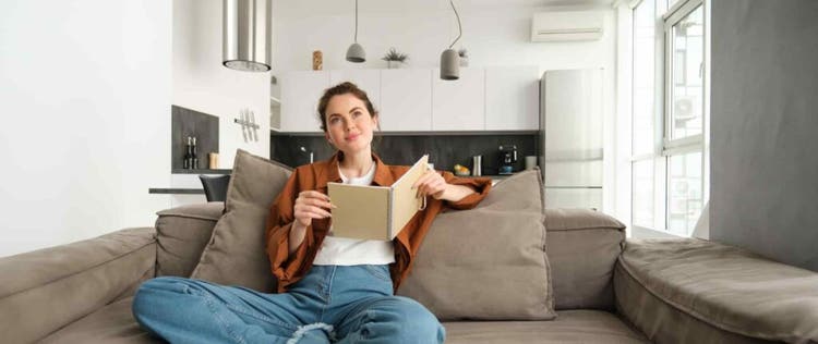 wanita duduk di sofa sambil membuka buku di dalam ruangan ber-ac